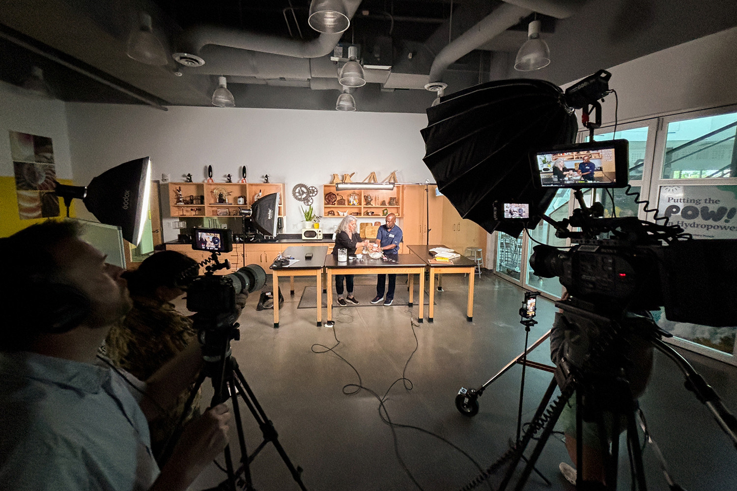 A behind-the-scenes shot showing lighting and several cameras recording a man and a woman in a kitchen studio. 